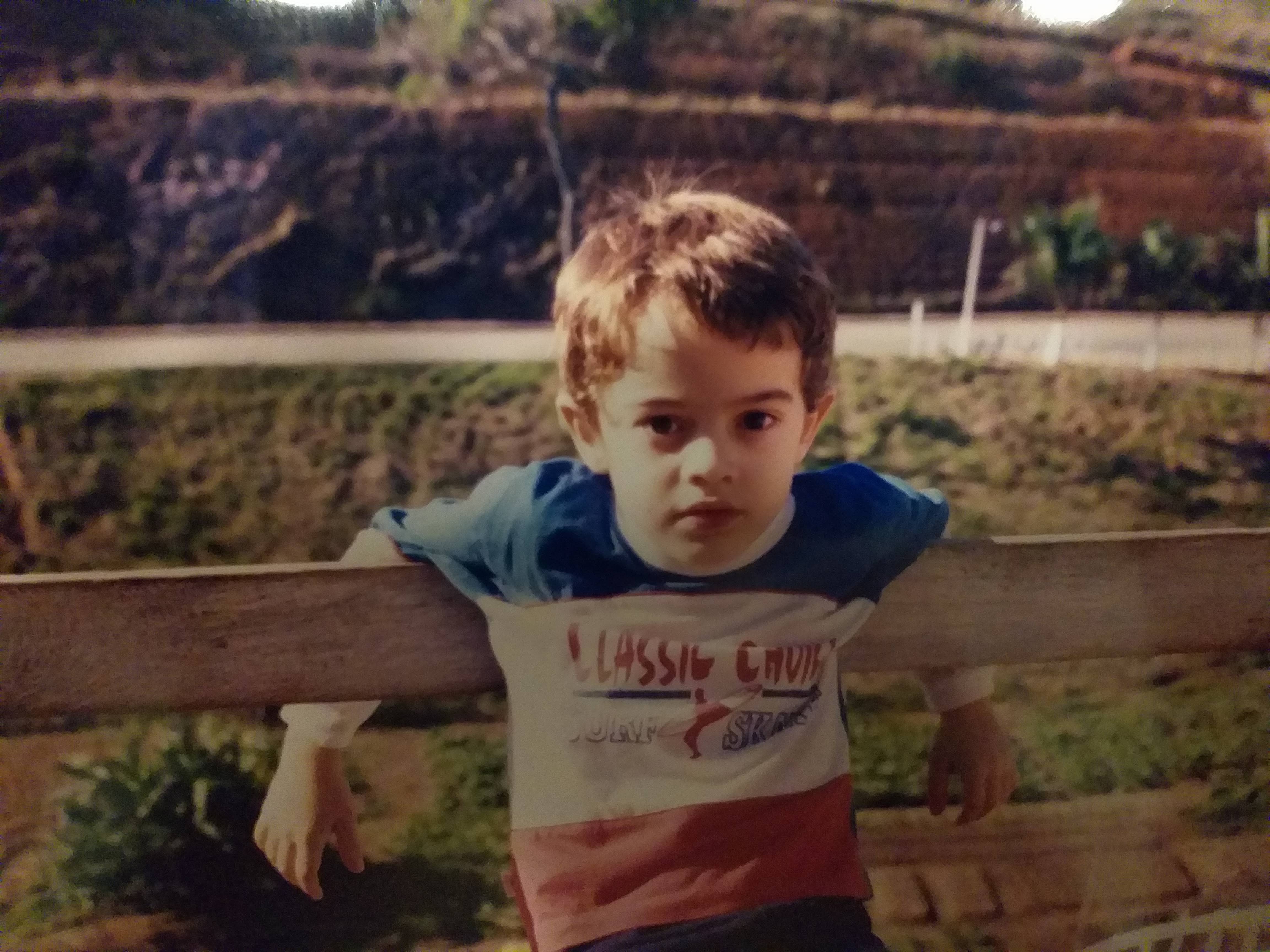A close-up, eye-level portrait of a young boy with wavy light brown hair and dark, expressive eyes looking directly at the camera with a neutral, calm expression. He is leaning his arms over a horizontal, light-colored wooden fence rail. The boy is wearing a color-blocked T-shirt consisting of a blue top section, a white middle section with the text 'CLASSIC CHOICE' in red and 'SURF SKATE' in blue alongside a small surfer graphic, and a red bottom section. He has long white sleeves visible underneath the T-shirt. The background is a slightly blurred outdoor setting featuring green grass, a dirt path, and a sloped, terraced hillside in the distance under warm, natural afternoon lighting, giving the photo a nostalgic, vintage feel